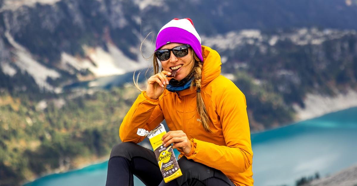 Woman on mountain smiles and takes a bite of a Zazubean Organic Chocolates bar